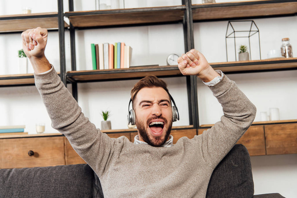 Exited man in headphones with hands in air on sofa in living room - Photo, Image