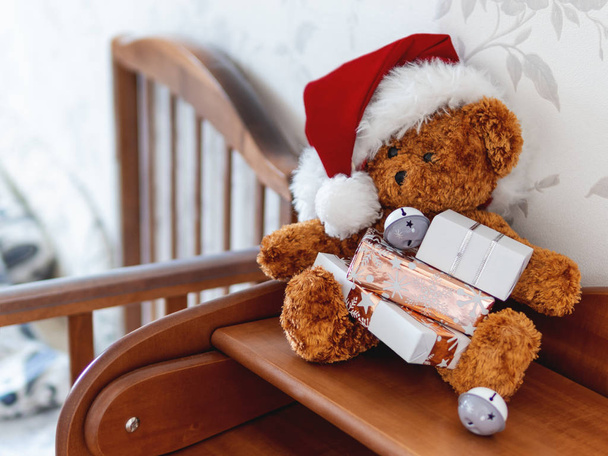 Teddy bear in Santa Claus hat. Plush toy with Christmas presents wrapped in white and golden paper. Kids room before New Year. - 写真・画像