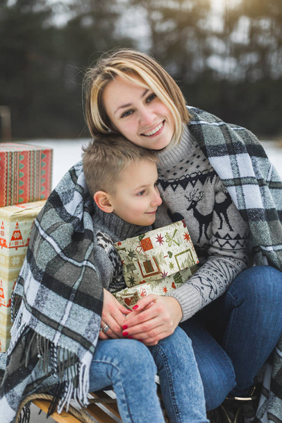 Mom and son enjoying snowy winter day outdoors, sitting on the sledge, hugging, covered with plaid and holding Christmas gifts - Фото, изображение