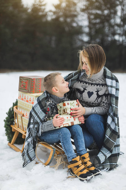 pretty young mother talk with child son while sitting on the wooden sledge outdoors in winter forest, covered with checkered plaid together. Family winter holidays outdoors. - Photo, image