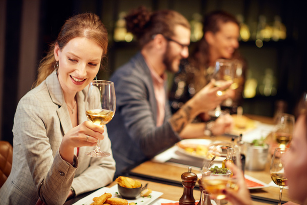 schöne kaukasische Brünette elegant gekleidet posiert mit einem Glas Wein, während sie mit ihren Freunden im Restaurant sitzt. - Foto, Bild