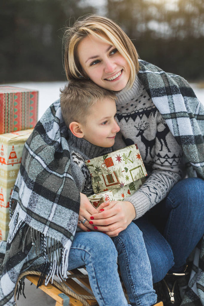 Mom and son enjoying snowy winter day outdoors, sitting on the sledge, hugging, covered with plaid and holding Christmas gifts - 写真・画像