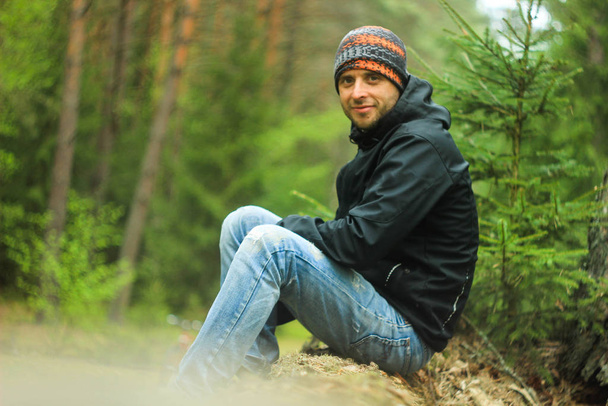 Mladý atraktivní muž turista s neoholeností ve sportovní bundě, modré džíny, buff a trekking boty sedí na kmeni stromu v podzimním lese. - Fotografie, Obrázek