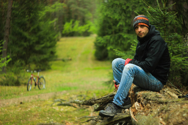 Jovem ciclista masculino atraente com unshaven em uma jaqueta esportiva, jeans azuis, um lustre e botas de trekking senta-se em um tronco de árvore na floresta de outono. No fundo é uma bicicleta
. - Foto, Imagem