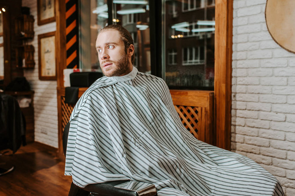 bearded man in stripped barbers cape sitting in barbershop  - Zdjęcie, obraz