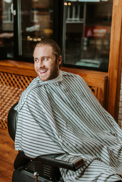 heureux homme barbu en stripped coiffeurs cape assis dans salon de coiffure
  - Photo, image