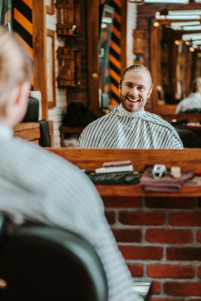 Selektiver Fokus eines glücklichen bärtigen Mannes, der im Friseurladen in den Spiegel schaut  - Foto, Bild