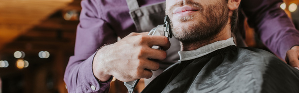 panoramic shot of barber holding trimmer while shaving bearded man  - Zdjęcie, obraz