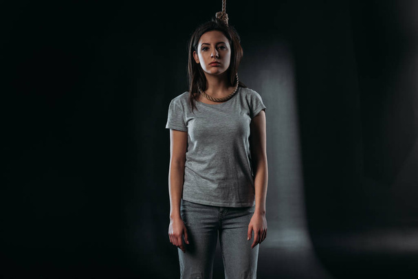 depressed young woman looking at camera while standing with noose on neck on black background - Photo, image