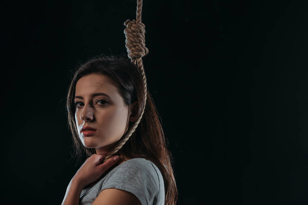 frustrated young woman looking at camera while standing with noose on neck isolated on black - Фото, изображение