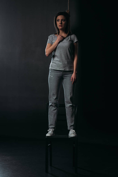 depressed woman standing on chair with noose on neck and looking away on black background - Photo, Image