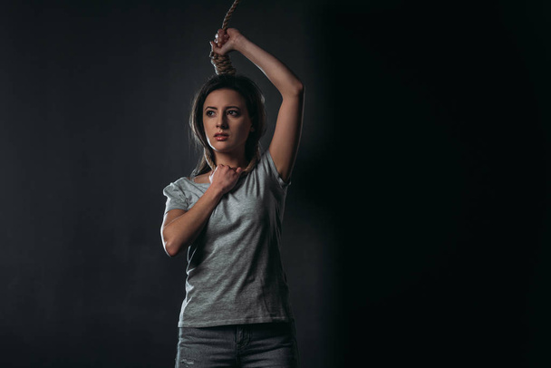 despaired woman looking away while standing with noose on neck on black background - Foto, immagini