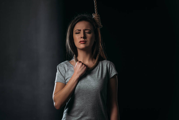 hopeless woman committing suicide while standing with noose on neck on black background - Fotografie, Obrázek
