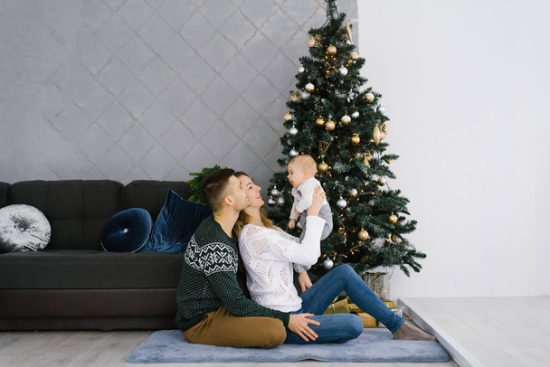 Egy apa és anya egy kisfiút tart egy karácsonyfa mellett. Karácsonyi dekoráció a belső tér a ház. A családi nyaralás fogalma. Boldog egy barátságos fiatal család. - Fotó, kép