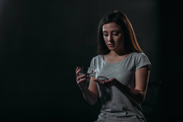 depressieve vrouw gaat zelfmoord te plegen terwijl het houden van handvol pillen en glas water geïsoleerd op zwart - Foto, afbeelding