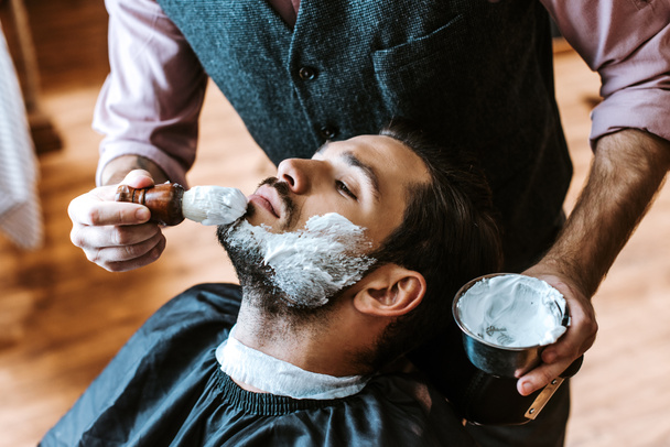 selektivní zaměření holiče nanášení holení krém na tvář člověka při držení misky  - Fotografie, Obrázek