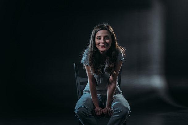 happy young woman smiling at camera while sitting in darkness on black background - Fotografie, Obrázek