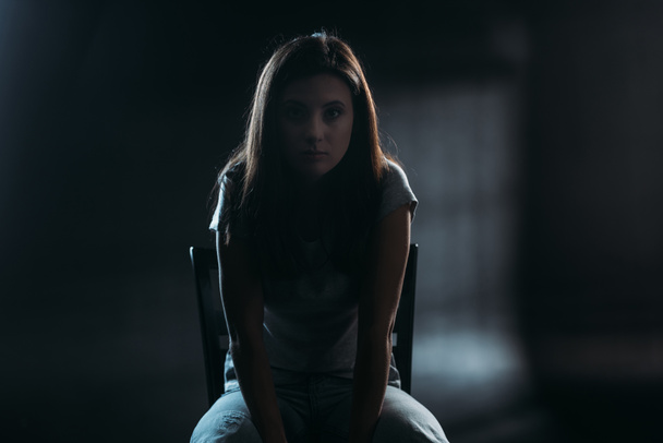 mujer joven sin esperanza mirando a la cámara mientras está sentado en la silla en la oscuridad sobre fondo negro
 - Foto, Imagen