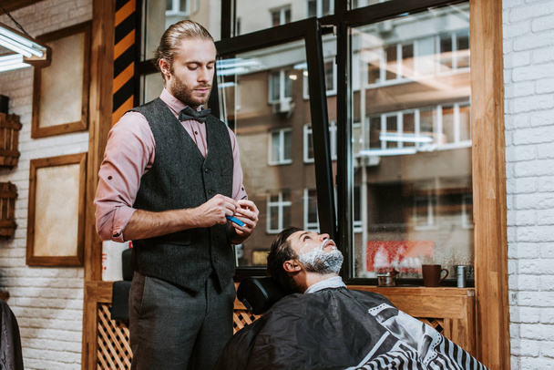 barbeiro bonito segurando navalha perto barbudo homem com creme de barbear no rosto
  - Foto, Imagem