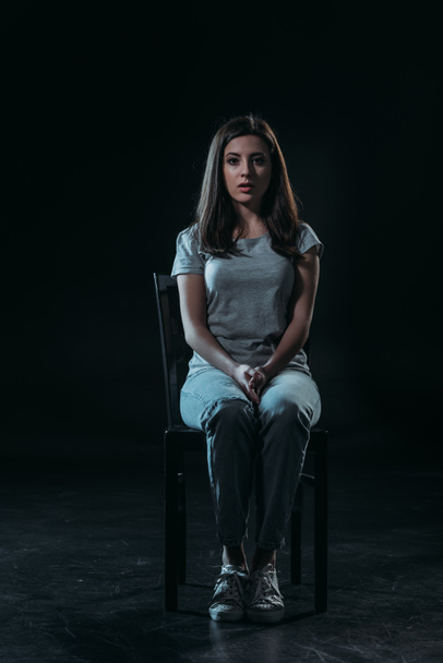 sad young woman looking at camera while sitting on chair in darkness on black background - Photo, Image