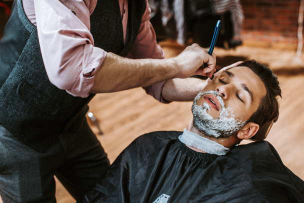 Friseur rasiert hübschen Mann mit Rasierschaum im Gesicht  - Foto, Bild