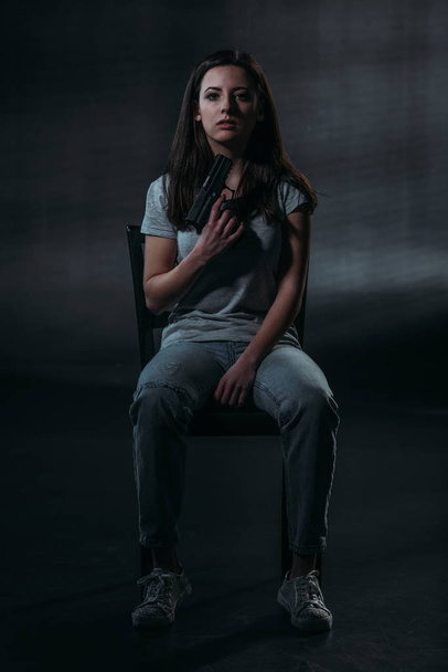 despaired woman holding gun near chin and looking at camera while sitting on chair on dark background  - Photo, image