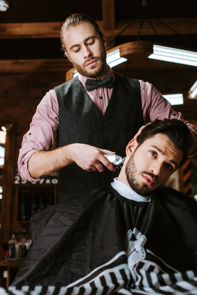 stylish barber holding trimmer while styling hair of handsome man  - Foto, imagen