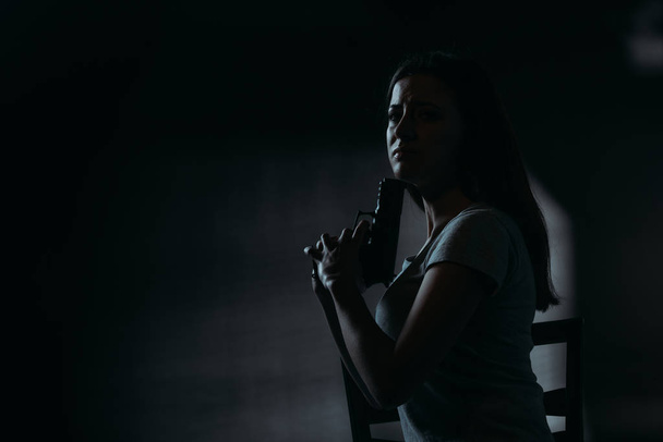 frustrated woman looking at camera while holding gun near chin on dark background  - Foto, imagen