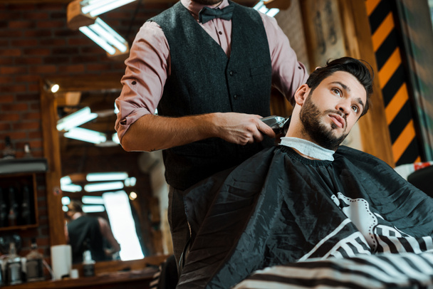 stylish barber holding trimmer while styling hair of man  - Photo, Image