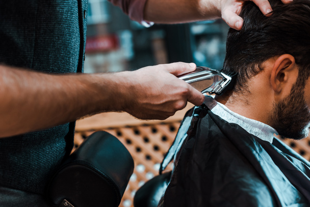 selective focus of barber holding trimmer while styling hair of man  - Foto, Bild