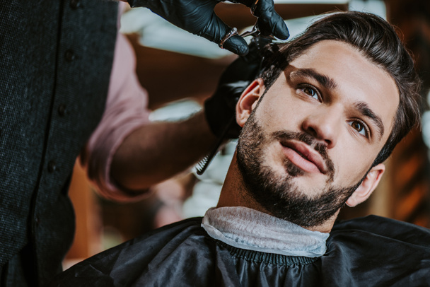 selektywne skupienie fryzjera w lateksowych rękawiczkach z nożyczkami podczas stylizacji włosów człowieka  - Zdjęcie, obraz