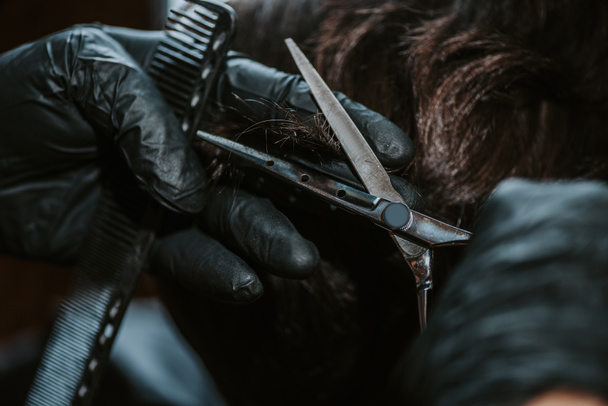 primo piano del barbiere in guanti di lattice che tengono pettine e forbici mentre acconciano i capelli dell'uomo
  - Foto, immagini