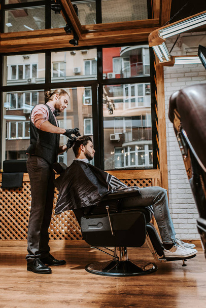 side view of handsome barber styling hair of man in modern barbershop  - Photo, Image