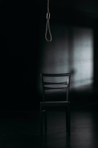 chair under hanging rope noose on black background with lighting, suicide prevention concept - Photo, image