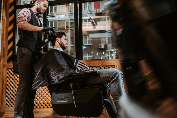 πλαϊνή άποψη του όμορφου κουρέα styling μαλλιά του γενειοφόρου άνδρα στο σύγχρονο κουρείο  - Φωτογραφία, εικόνα