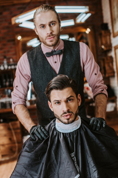 el barbero guapo en los guantes de látex poniendo las manos sobre los hombros del hombre en la barbería
  - Foto, imagen