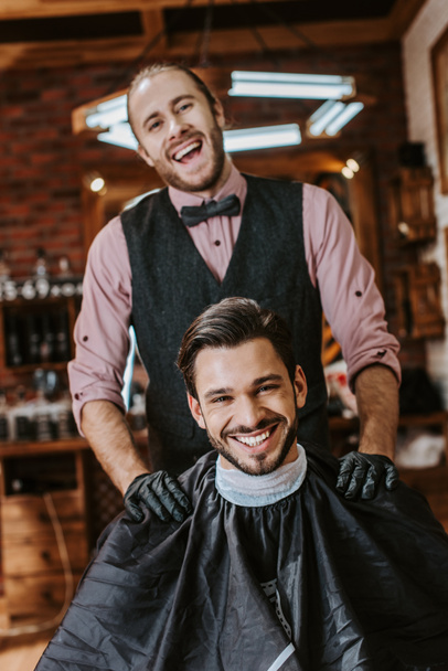 handsome barber in latex gloves putting hands on shoulders of happy man in barbershop  - Fotó, kép