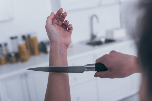 partial view of depressed woman committing suicide by cutting veins with knife in kitchen - Foto, afbeelding
