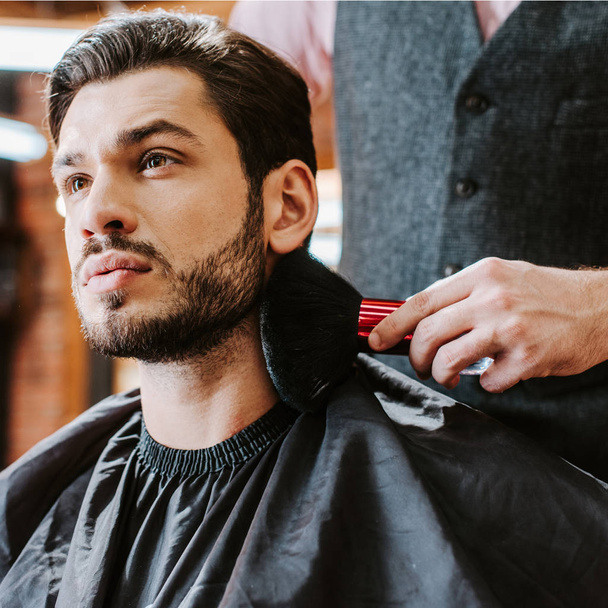selective focus of barber holding cosmetic brush near face of handsome bearded man  - Fotoğraf, Görsel