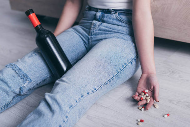 cropped view of unconscious woman sitting on floor with bottle of alcohol and handful of pills - Foto, immagini