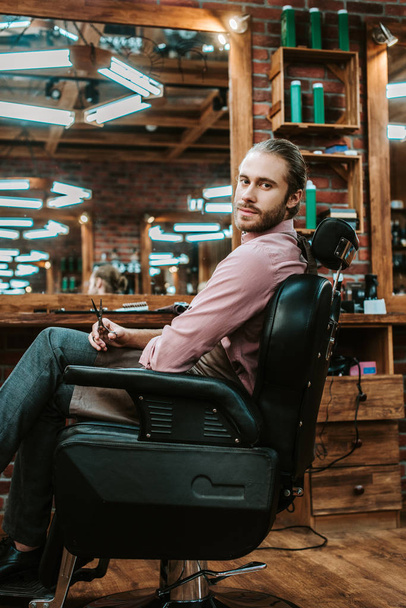barbero guapo y barbudo mirando a la cámara mientras está sentado en el sillón
  - Foto, Imagen