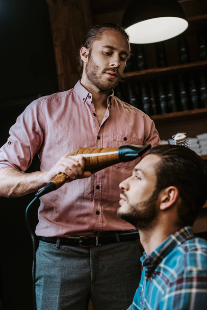 przystojny fryzjer gospodarstwa suszarka do włosów w pobliżu brodaty człowiek  - Zdjęcie, obraz