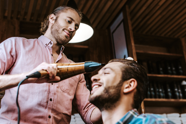 foco seletivo de barbeiro bonito segurando secador de cabelo perto de homem feliz
  - Foto, Imagem