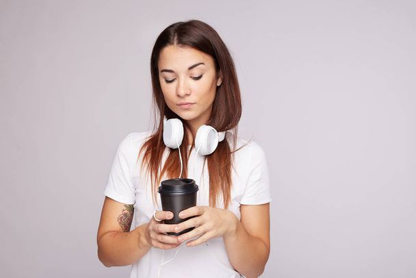 Indoor shot of timid female teenage looking down with dreamy thoughtful serious face expression, has long straight dark hair, holds coffee cup, dressed in casual white t shirt, isolated on grey wall. - Photo, image