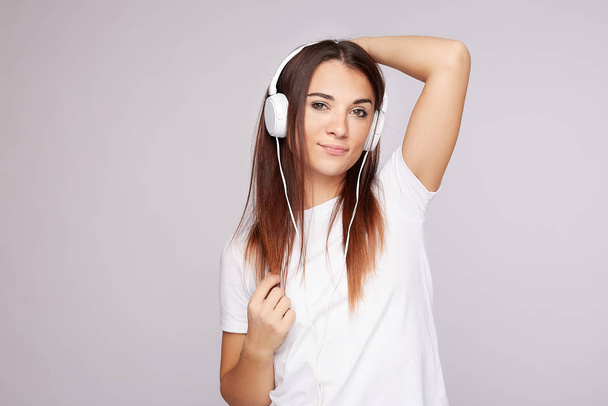 Snímek hlavy mladé příjemné ženy, jak poslouchá, užívá si hudby přes online přehrávač, nosí bílé sluchátka, tričko, pózuje uvnitř u šedé stěny studia s kopírovacím prostorem pro text nebo reklamní obsah - Fotografie, Obrázek