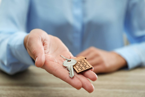 Real estate agent holding house key with trinket at wooden table, closeup - 写真・画像