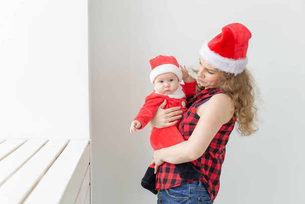 Family, childhood and Christmas concept - Young mother holding baby in santa suit indoor - Fotó, kép