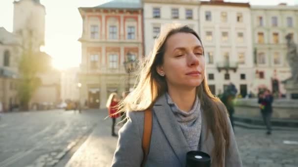 Woman with a thermos cup in hand walking down the street and admires the architecture of the old city at sunset on a cold autumn day - Video