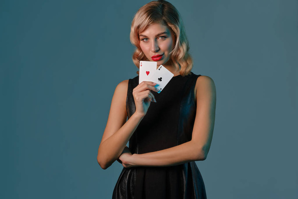 Blonde girl in black stylish dress showing two playing cards, posing against blue background. Gambling entertainment, poker, casino. Close-up. - Valokuva, kuva
