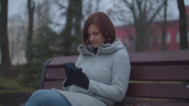 Barna nő felnőtt okostelefonnal ül padon őszi parkban. Nő mosolyog, miközben szörfözés online telefonon.  - Felvétel, videó